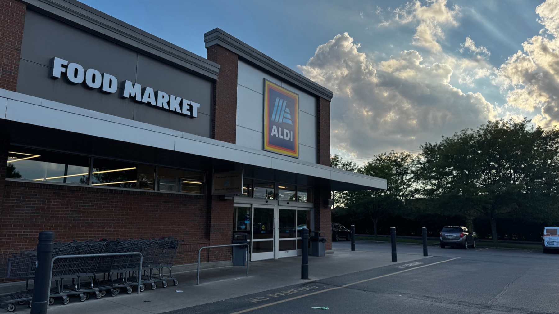 An ALDI storefront at dusk, its lit FOOD MARKET sign and blue logo above the entrance and carts lined up outside